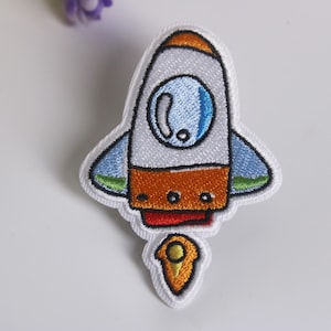 Könnte beinhalten: Ein gestickter Bügelaufkleber mit einem Cartoon-Raketenfahrzeug mit Flamme in Weiß, Orange, Blau und Grün.