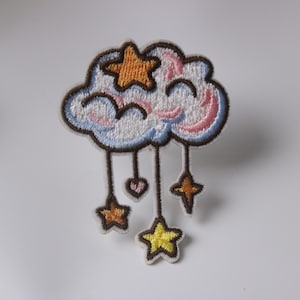 May include: Embroidered patch featuring a cloud with a white base, outlined in brown. The cloud has pink and blue accents. Hanging from the cloud are brown strings with a heart and stars in orange and yellow. This patch is ideal for decorating clothing or accessories.