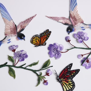 Puede incluir: Parches bordados con pájaros de alas azules y rosas, mariposas y flores moradas sobre ramas verdes. Las mariposas tienen diseños naranjas, rojos y amarillos. Los parches están sobre un fondo blanco.