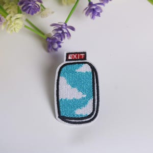 May include: An embroidered iron-on patch with a blue and white design of a bottle with clouds inside. The bottle has a red label that says "EXIT".