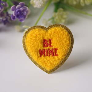 Puede incluir: Un parche amarillo en forma de corazón con las palabras "BE MINE" bordadas en rojo. El parche tiene un borde dorado brillante. Fondo blanco con flores moradas y blancas.