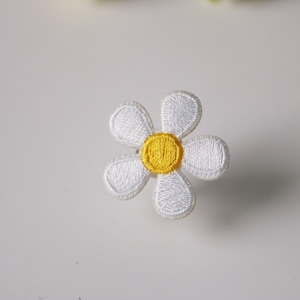 Puede incluir: Un parche de flor bordado de color blanco con un centro amarillo. La flor tiene seis pétalos.