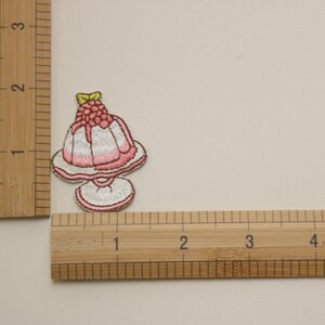 Peut inclure: Un patch brod&eacute; thermocollant repr&eacute;sentant un dessert rose et blanc sur une assiette avec une framboise sur le dessus. Le patch mesure environ 5 cm de large.