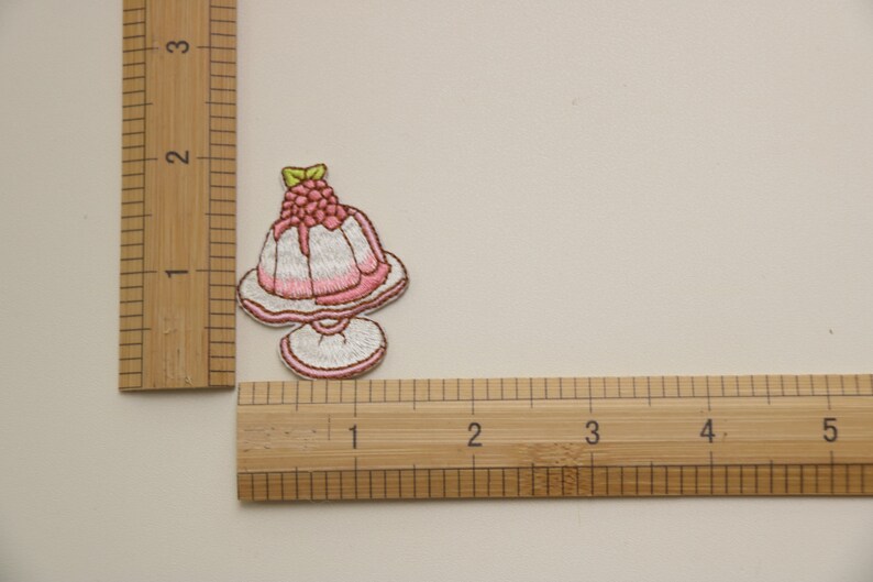 Peut inclure: Un patch brod&eacute; thermocollant repr&eacute;sentant un dessert rose et blanc sur une assiette avec une framboise sur le dessus. Le patch mesure environ 5 cm de large.