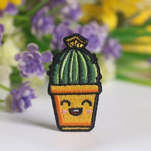 Puede incluir: Parche bordado con un cactus sonriente en una maceta amarilla. El cactus es verde con líneas verticales, y la maceta es amarilla con un contorno negro. El parche tiene un diseño alegre, tipo caricatura, ideal para ropa o accesorios.