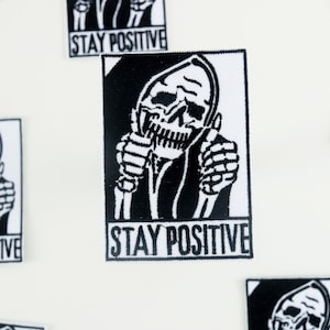 May include: Black and white embroidered patch featuring a skeleton giving a thumbs up with the text "STAY POSITIVE" below.