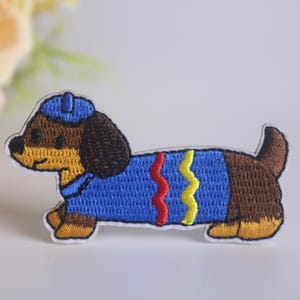 Könnte beinhalten: Gestickter Bügelaufkleber mit einem braunen Dackel, der einen blauen Pullover mit roten und gelben Akzenten und eine blaue Mütze trägt.