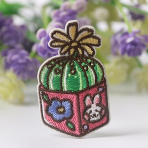 Pode incluir: Etiqueta bordada para passar a ferro com um cacto verde com uma flor amarela no topo, num vaso rosa com uma flor azul e um coelho branco no lado.