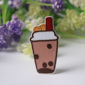 Könnte beinhalten: Gestickter Aufnäher mit einem braunen Bubble Tea-Getränk mit weißem Rand, braunen Boba-Perlen und einem roten Strohhalm. Der Aufnäher hat ein detailliertes Design mit braunen Umrissen. Der Hintergrund umfasst lila und grüne Blumen.