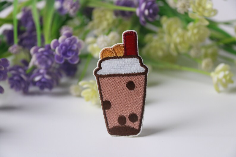 Może przedstawiać: Haftowana naszywka przedstawiająca brązowy nap&oacute;j bubble tea z białym obramowaniem, brązowymi perełkami boba i czerwoną słomką. Naszywka ma szczeg&oacute;łowy wz&oacute;r z brązowym obrysem. Tło obejmuje fioletowe i zielone kwiaty.