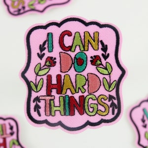 Może przedstawiać: Różowa haftowana naszywka do prasowania z tekstem "I can do hard things" w kolorowych literach otoczonych zielonymi liśćmi i czerwonymi kwiatami.