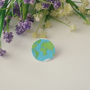 Pode incluir: Um remendo bordado azul e verde da Terra sobre um fundo branco. O remendo &eacute; cercado por flores roxas e brancas.