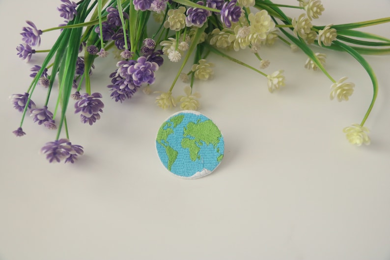 Pode incluir: Um remendo bordado azul e verde da Terra sobre um fundo branco. O remendo &eacute; cercado por flores roxas e brancas.
