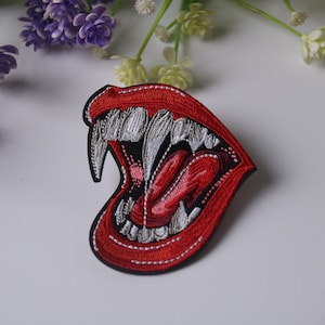 Puede incluir: Parche bordado rojo de una boca grande abierta con dientes blancos afilados y una lengua roja.