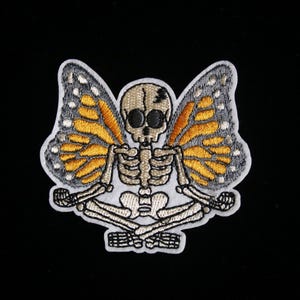 Op de afbeelding: Geborduurde patch met een skelet met monarchvlindervleugels. Het skelet zit in een zittende yogahouding, met oranje, gele en grijze vleugels. De patch staat op een zwarte achtergrond.