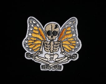 May include: Embroidered patch of a skeleton with monarch butterfly wings. The skeleton is in a seated yoga pose, with orange, yellow, and grey wings. The patch is set against a black background.