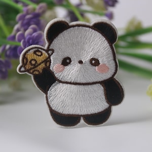 Może przedstawiać: Naszywka haftowana do prasowania przedstawiająca uroczego kreskowego pandę trzymającego planetę. Panda jest biały z czarnymi uszami, oczami i łapami. Planeta jest żółta z brązowym pierścieniem.