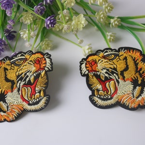 以下が含まれることがあります： 口を開けた虎の頭が描かれた、アイロン接着式の刺繍パッチ2枚。黄色とオレンジの毛並み、黒い縞模様、白いアクセントが特徴です。