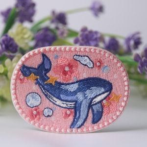 Puede incluir: Un parche bordado rosa con una ballena azul nadando a través de un campo de flores rosas y nubes blancas. La ballena tiene una barriga blanca y está rodeada de burbujas blancas.