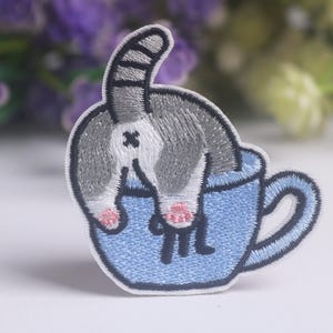 Op de afbeelding: Een geborduurde patch met een grijs-witte cyperse kat die met de achterkant omhoog in een lichtblauwe theekop zit. De kat heeft een gestreepte staart, roze pootkussentjes en de theekop heeft een donkerblauw oortje en een donkere rand.