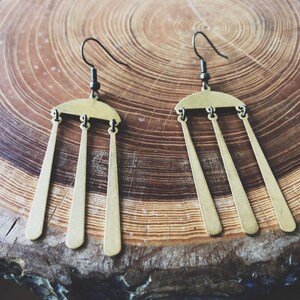 Peut inclure: Une paire de boucles d'oreilles couleur laiton au design unique. Chaque boucle d'oreille pr&eacute;sente un dessus incurv&eacute; avec trois pendentifs allong&eacute;s en forme de trap&egrave;ze. Les boucles d'oreilles ont des crochets noirs et sont pr&eacute;sent&eacute;es sur une surface en bois.