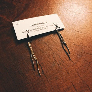 May include: A pair of gold-toned earrings with a long, thin, and slightly wavy design. The earrings are displayed on a white card with the word "contentious" written on it. The card also includes the definition of the word, "(kan-ten shas) They are so contentious that we simply can't reason with them."