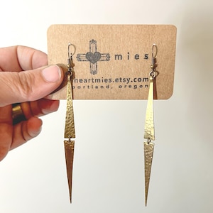 May include: A pair of long, brass-colored earrings with a hammered texture. Each earring has a triangular shape with a pointed end, connected by a small rectangular piece. The earrings are displayed on a brown card with the text "heartmies.etsy.com portland, oregon".