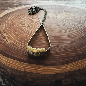 Puede incluir: Un collar de oro con un diseño único que presenta una serie de anillos apilados y un colgante largo y delgado. El collar se muestra sobre una superficie de madera.