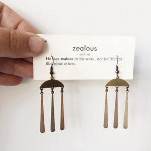 Peut inclure: Une paire de boucles d'oreilles en laiton avec un dessus en demi-cercle et trois triangles allong&eacute;s suspendus. Les boucles d'oreilles sont pr&eacute;sent&eacute;es avec une carte qui dit "zealous" avec une d&eacute;finition. Sur fond blanc.