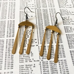 Peut inclure: Boucles d'oreilles en m&eacute;tal dor&eacute;, en forme de parapluie sur le dessus avec trois rectangles pendants. Les boucles d'oreilles sont pr&eacute;sent&eacute;es sur un fond de papier vintage avec du texte imprim&eacute;.