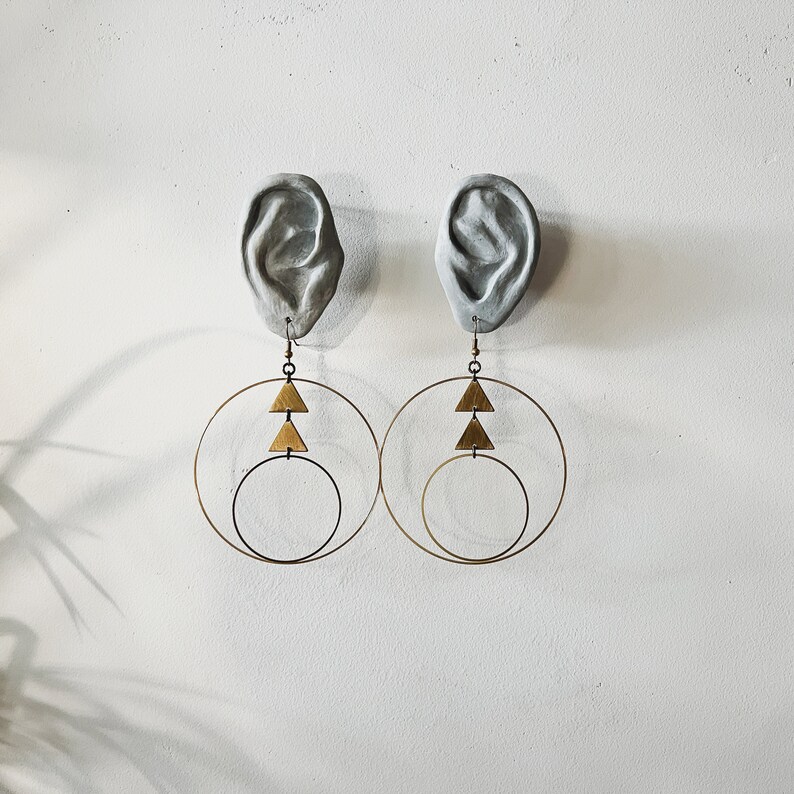 Peut inclure: Une paire de boucles d'oreilles cr&eacute;oles dor&eacute;es avec un motif triangulaire. Les boucles d'oreilles sont suspendues &agrave; un pl&acirc;tre d'oreille.