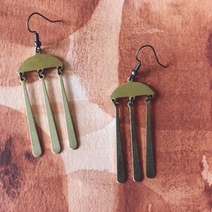 Peut inclure: Une paire de boucles d'oreilles couleur laiton. Chaque boucle d'oreille pr&eacute;sente un dessus en demi-cercle avec trois pendules rectangulaires verticaux et allong&eacute;s. Les crochets d'oreilles sont gris fonc&eacute;. Le fond est de couleur brun-rouge&acirc;tre mouchet&eacute;.
