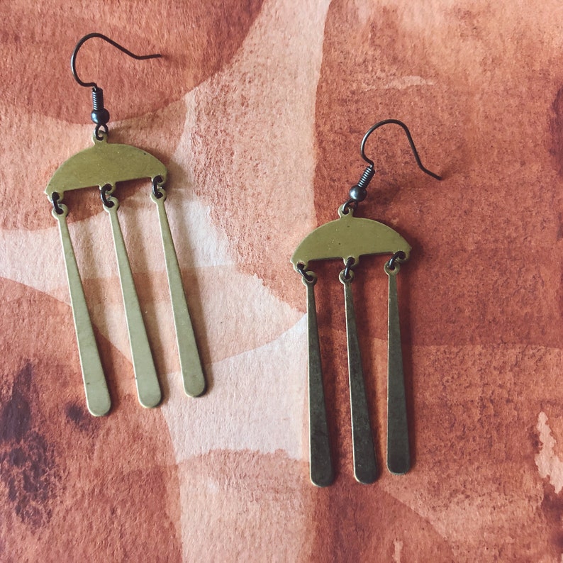 Peut inclure: Une paire de boucles d'oreilles couleur laiton. Chaque boucle d'oreille pr&eacute;sente un dessus en demi-cercle avec trois pendules rectangulaires verticaux et allong&eacute;s. Les crochets d'oreilles sont gris fonc&eacute;. Le fond est de couleur brun-rouge&acirc;tre mouchet&eacute;.