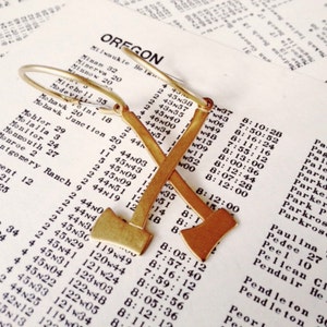 May include: A pair of gold-toned earrings shaped like crossed axes. The earrings are hanging from a piece of paper with text on it.