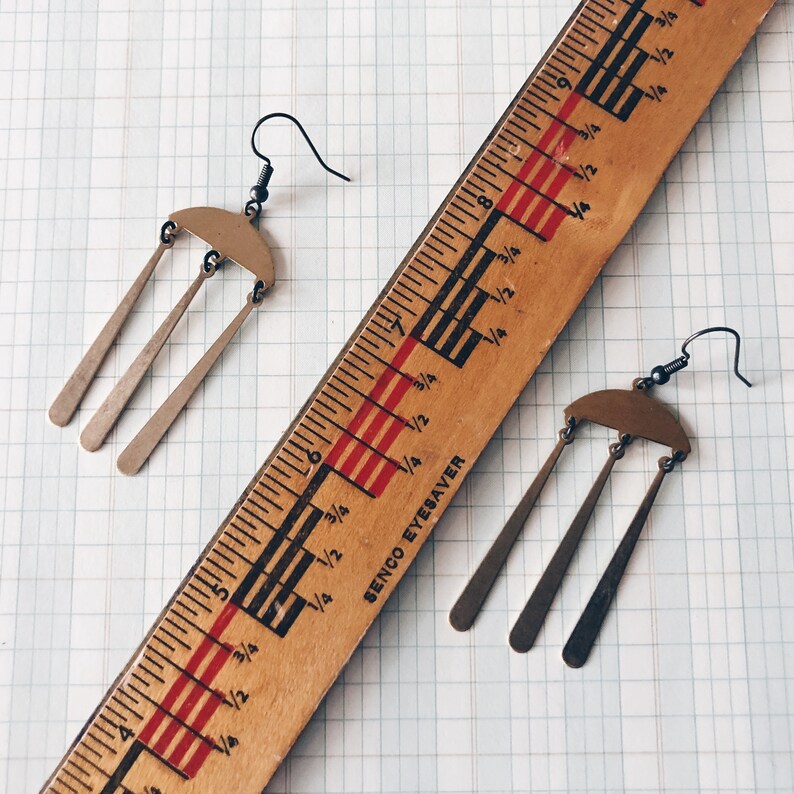 Peut inclure: Boucles d'oreilles de couleur bronze, compos&eacute;es d'une partie sup&eacute;rieure en demi-cercle et de trois pendants allong&eacute;s. Une r&egrave;gle en bois vintage avec des marquages noirs et rouges est &agrave; c&ocirc;t&eacute;. Les mesures sont en centim&egrave;tres.