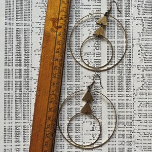 Peut inclure: Une paire de boucles d'oreilles cr&eacute;oles dor&eacute;es avec un pendentif triangulaire. Les boucles d'oreilles sont sur un fond blanc avec une r&egrave;gle.