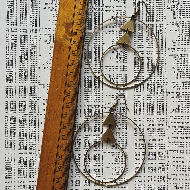 Peut inclure: Une paire de boucles d'oreilles cr&eacute;oles dor&eacute;es avec un pendentif triangulaire. Les boucles d'oreilles sont sur un fond blanc avec une r&egrave;gle.