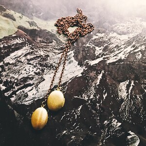 May include: A long gold chain necklace with two round gold locket pendants. The necklace is photographed against a backdrop of a mountain range.