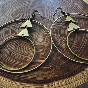 Peut inclure: Une paire de boucles d'oreilles cr&eacute;oles dor&eacute;es avec deux formes triangulaires suspendues &agrave; chaque cr&eacute;ole. Les boucles d'oreilles sont sur une surface en bois.