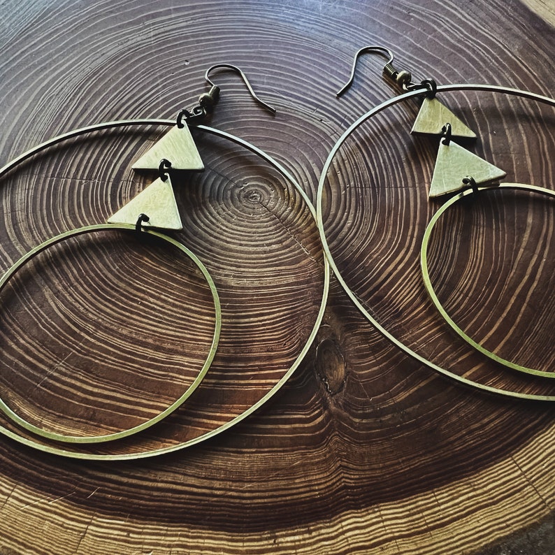 Peut inclure: Une paire de boucles d'oreilles cr&eacute;oles dor&eacute;es avec deux formes triangulaires suspendues &agrave; chaque cr&eacute;ole. Les boucles d'oreilles sont sur une surface en bois.