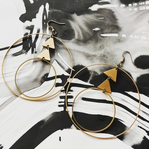Peut inclure: Une paire de boucles d'oreilles cr&eacute;oles dor&eacute;es avec deux formes triangulaires suspendues &agrave; chaque cr&eacute;ole. Les boucles d'oreilles sont sur un fond blanc et noir.