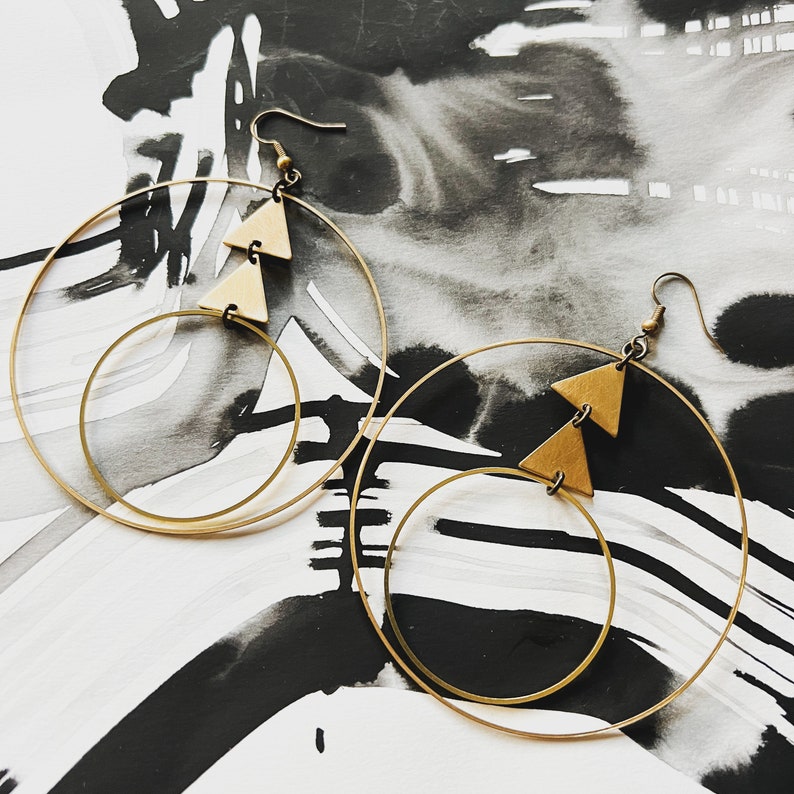 Peut inclure: Une paire de boucles d'oreilles cr&eacute;oles dor&eacute;es avec deux formes triangulaires suspendues &agrave; chaque cr&eacute;ole. Les boucles d'oreilles sont sur un fond blanc et noir.