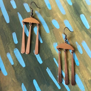 Peut inclure: Une paire de boucles d'oreilles couleur cuivre. Chaque boucle d'oreille pr&eacute;sente un dessus en demi-cercle avec trois trap&egrave;zes allong&eacute;s pendants. Les boucles d'oreilles sont suspendues &agrave; des crochets d'oreille noirs. Le fond est un m&eacute;lange de coups de pinceau verts et bleus.
