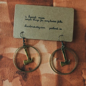 Può includere: Orecchini a cerchio con ciondoli a forma di ascia dorata. Gli orecchini sono appesi a ganci neri. Sullo sfondo, un'etichetta marrone con il testo "i heart mies" e "iheartmies.etsy.com".