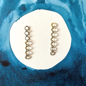 May include: Two pairs of gold-toned chain earrings. Each earring features a series of small, connected circles. The earrings are displayed on a white surface against a backdrop of blue watercolor paint.