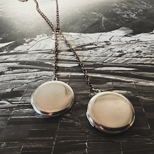 May include: Two silver locket necklaces with a chain made of copper-toned metal. The lockets are round and have a smooth, shiny surface. The necklaces are hanging against a backdrop of a black and white aerial photograph.