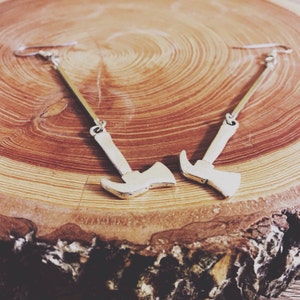 May include: A pair of silver axe earrings hanging from a wooden surface. The earrings are long and dangle, with the axe head at the bottom.