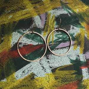 May include: Two copper-colored hoop earrings. The earrings are circular and have a simple, minimalist design. The background is a colorful abstract painting with green, yellow, red, and purple brushstrokes.
