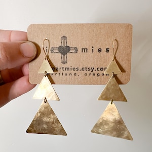 Puede incluir: Pendientes de triángulo de oro con textura martillada. Los pendientes están superpuestos en un diseño en cascada. Los pendientes se muestran en una tarjeta marrón con el texto "mies" y "heartmies.etsy.com" impreso en ella.