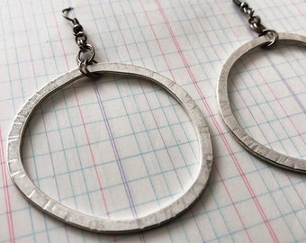 Puede incluir: Un par de pendientes de aro de plata con textura martillada. Los pendientes son grandes y tienen un diseño simple y moderno.
